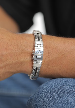 Silver bracelet featuring braided metal accents, rectangular clasp with logo, textured surface, and sleek design. Worn on a wrist.