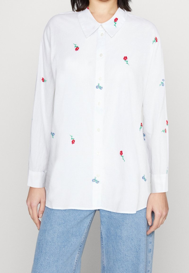 Chemise blanche boutonnée avec broderie florale colorée. Manches longues avec poignets, coupe décontractée, et texture douce.
