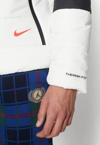 Vit puffjacka med svarta detaljer, utrustad med dragkedja och texten "THERMA-FIT" på mudden, kombinerad med rutiga byxor i blått och grönt.