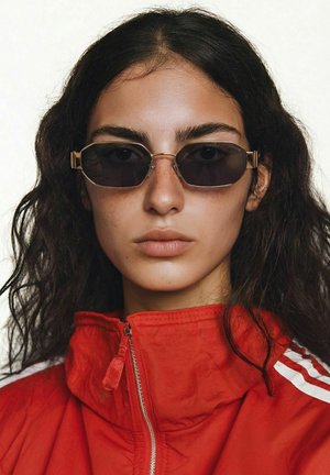 Femme aux cheveux bouclés foncés portant des lunettes de soleil rectangulaires à monture dorée et une veste zippée rouge avec des bandes blanches sur les épaules.