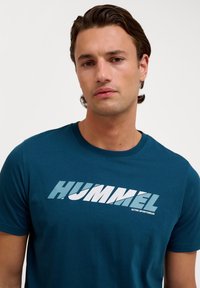 Turkos bomullströja med en stor "HUMMEL" grafik i vitt och ljusblått. Klassisk rundhals och korta ärmar.