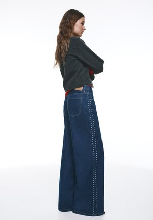 Džínsy široké na nohách v tmavomodrej farbe s hladkým textúrou. Majú strieborné cvoky po bočnom šve. Vysoký pás.