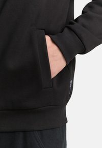 Schwarzer Sweatshirt mit geripptem Muster, ausgestattet mit einer Seitentasche und Bündchen an den Ärmeln. Kleines Markenetikett an der unteren Seite sichtbar.