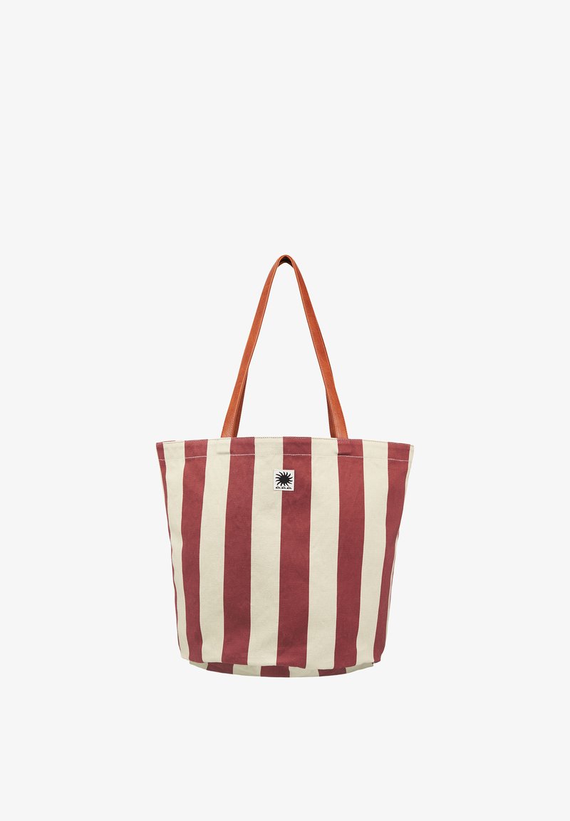 Sac fourre-tout en toile avec des rayures verticales rouges et blanches et des poignées en cuir marron, arborant un petit emblème noir en forme de soleil à l'avant.