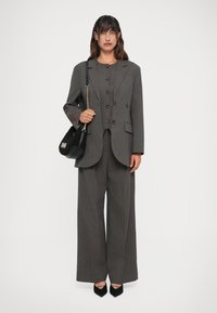 Sieviete valkā pelēku svītrainu oversize bleizeri un atbilstošas platām biksēm, tur rokās melnu rokassomu ar zelta ķēdes siksnu.