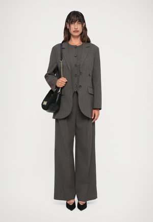 Žena u sivoj oversized blazerici s prugicama i širokim hlačama od istog materijala, drži crnu torbu s zlatnim lančićem.