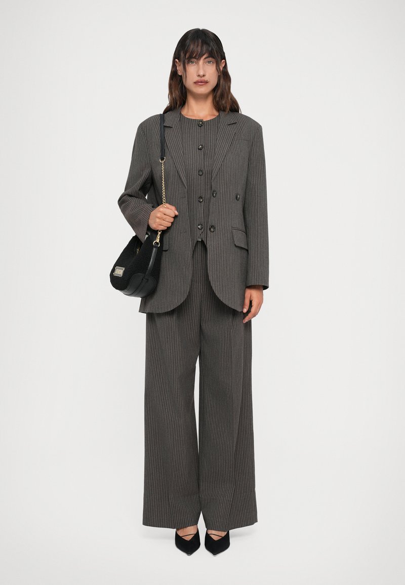 Sieviete valkā pelēku svītrainu oversize bleizeri un atbilstošas platām biksēm, tur rokās melnu rokassomu ar zelta ķēdes siksnu.