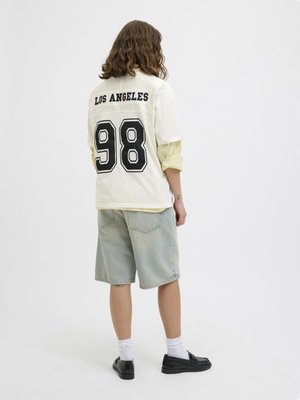 Vállig érő hajú személy fehér "Los Angeles 98" sportmezben, világos farmer rövidnadrágban, fehér zokniban és fekete cipőben, háttal áll.