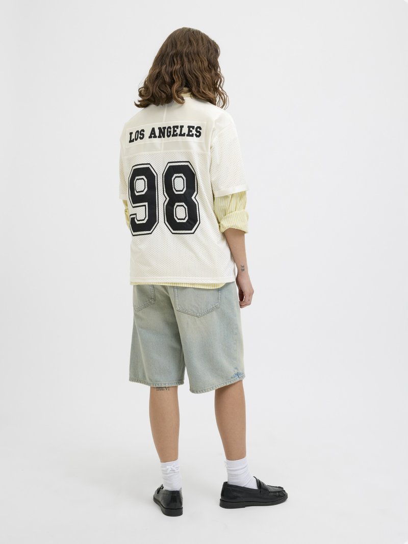 Personne aux cheveux mi-longs portant un maillot de sport blanc "Los Angeles 98", un short en jean clair, des chaussettes blanches et des chaussures noires, de dos.