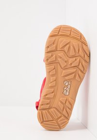 Červené sandály s texturovanou gumovou podrážkou a hlubokými drážkami pro lepší přilnavost. Logo "Jack Wolfskin" viditelné na patě. Nastavitelný pásek.