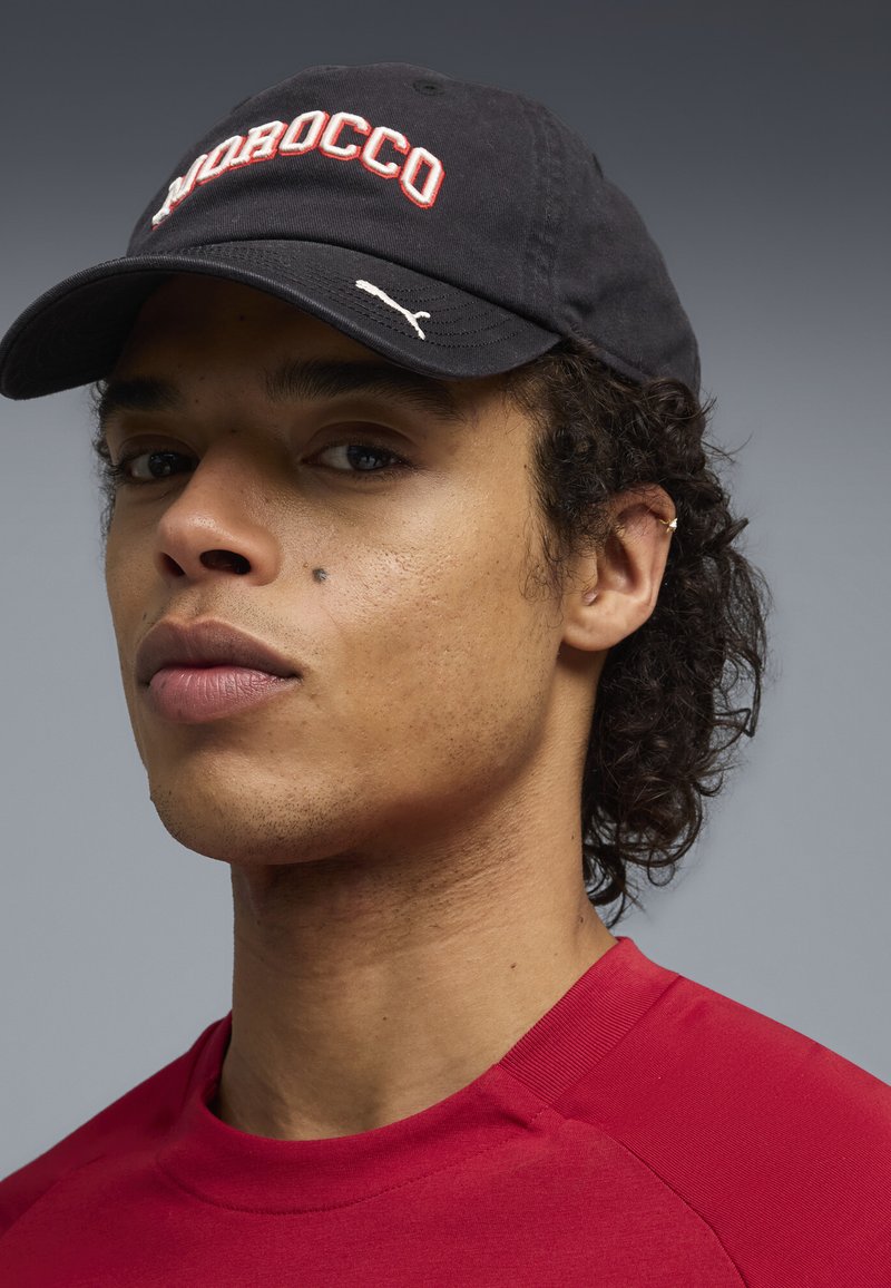 Jeune homme aux cheveux bouclés portant une casquette noire portant l'inscription « Morocco » et une chemise rouge, tourné légèrement vers la gauche sur un fond gris.