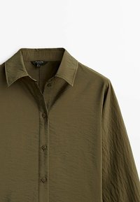 Olivgrön skjorta med krage, långa ärmar, texturerad tyg och synlig söm längs knäppningen och manschetterna.
