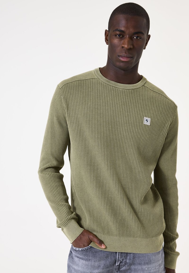 Olivgrüner, strukturierter Langarmpullover mit gerippten Bündchen und Saum, ausgestattet mit einem kleinen Logopatch auf der Brust. Getragen mit grauer Jeans.