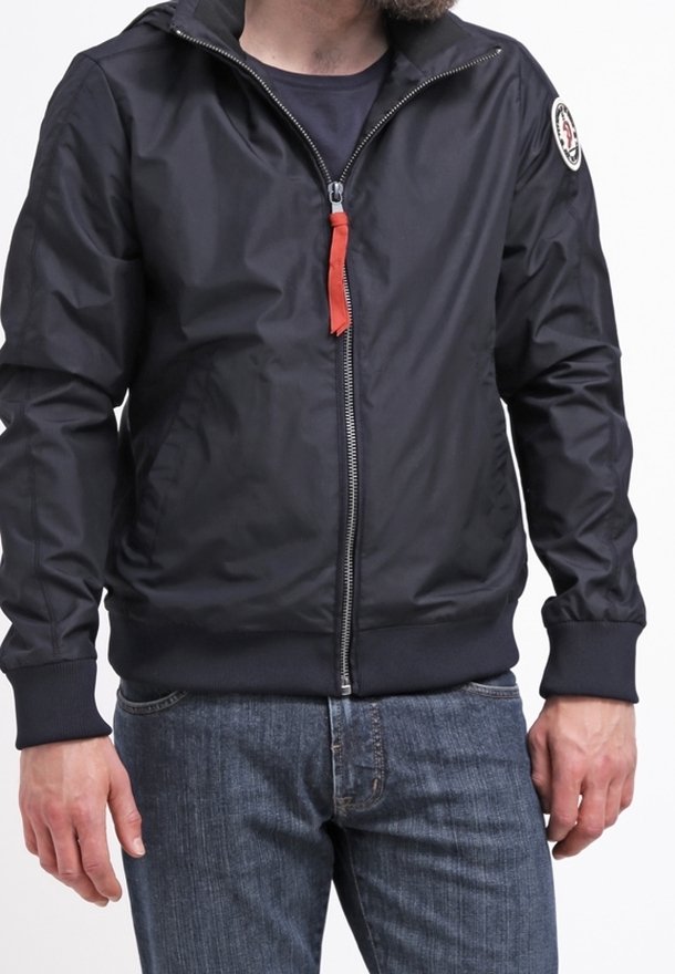 Homme portant une veste noire zippée avec une tirette rouge et un patch sur la manche, associée à un jean bleu.