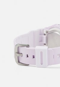 Correa de reloj de goma morada con herrajes de metal plateado, que presenta una textura suave y orificios ajustables para el tamaño. Acentuada con un sujetador blanco.