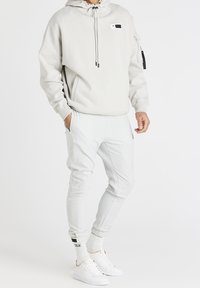 Lys grå hættetrøje med snøre i hætten, forstærket lomme foran og sidelommer med lynlås. Kombineret med matchende grå joggingbukser og hvide sneakers.