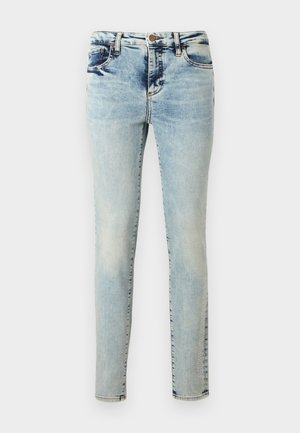 Blugi skinny albastru deschis cu spălare estompată, buzunare frontale, închidere cu nasturi și logo subtil pe lateral aproape de tiv, pe fundal alb.