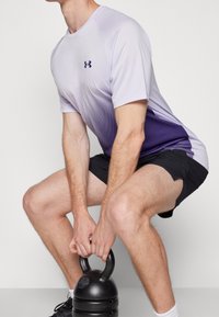 Camiseta deportiva morada y blanca con puntos en degradado, de manga corta, combinada con pantalones cortos negros. Persona sosteniendo una kettlebell negra.