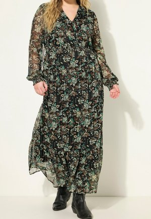 Femme portant une longue robe noire à fleurs avec des manches longues transparentes et des bottines noires, debout devant un fond clair uni.