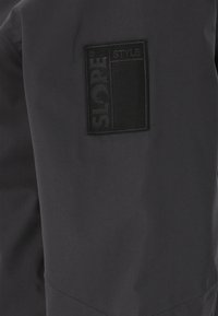 Svart jacka med en texturerad lappp som har texten "SLOPE STYLE"; slät material och snedställda sömmar synliga i designen.