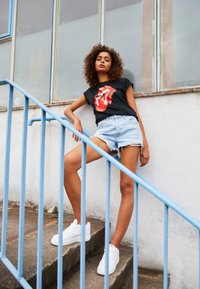 Sort T-shirt med rødt Rolling Stones-logo, denimshorts med frynser og hvide sneakers. Blå metalgelænder og betontrapper.