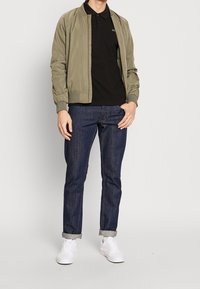Olivově zelená bomber bunda přes černé polokošile, v kombinaci s tmavě modrými džínami a bílými sportovními teniskami. Jednoduchý design, volný střih.