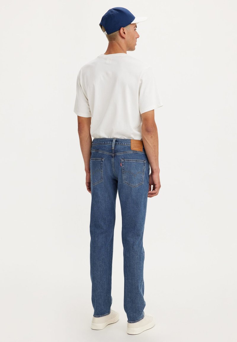 Jean en denim bleu moyen avec une coupe droite, présentant une patch en cuir brun sur la taille arrière et des détails d'usure subtile.