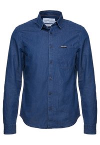 Mörkblå denimskjorta med knäppning nedtill, långa ärmar och en ficka på bröstet. Har en logotag på fickan.