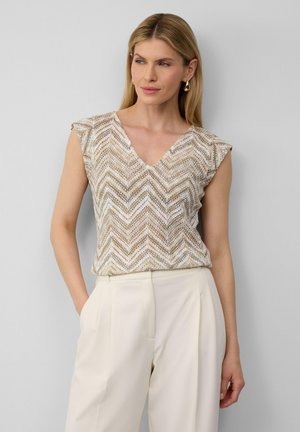 Beige gemusterte Bluse mit V-Ausschnitt und kurzen Ärmeln, die ein Zickzackdesign und transparente Textur aufweist, kombiniert mit hellen Hosen.