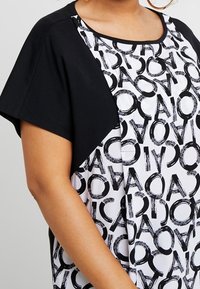 Černobílý top s kulatým výstřihem a krátkými rukávy. Černé rukávy kontrastují s tělem vzorovaným výraznými, překrývajícími se písmeny.