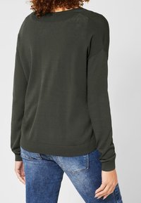 Tmavě zelený svetr s dlouhými rukávy, žebrovanými manžetami a kulatým výstřihem, v kombinaci s modrými džíny. Měkký, texturovaný materiál, pohodlný design.