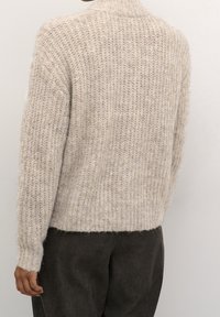 Suéter de punto de un beis claro con un cuello alto, textura acanalada, corte holgado y mangas anchas, combinado con pantalones oscuros y texturizados.