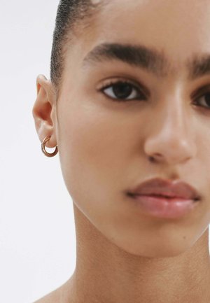 Pendiente de aro dorado exhibido en la oreja izquierda. Diseño circular y suave con un acabado brillante, complementado por el tono de piel neutro del modelo.