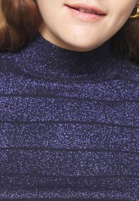 Violetti, ribbineulottu poolopaita, jossa on kimaltava tekstuuri ja vaakasuorat raidat. Malli on vartalonmyötäinen ja korostaa kaula-aukkoa.