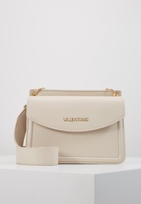 Beige läderhandväska med klaffstängning, guldfärgade beslag, präglad logotyp och avtagbar rem. Har rena linjer och strukturerad design.