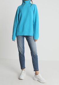 Pull à col roulé côtelé turquoise vif avec des manches longues, associé à un jean ajusté bleu clair et des baskets blanches. Design simple et moderne.