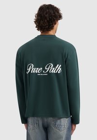 Donkergroene, lange mouwen shirt van gladde stof met de tekst "Pure Path" en "OWN THE JOURNEY" gedrukt in het wit op de achterkant.