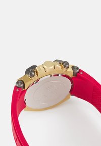 Rote Silikonarmbanduhr mit einem vergoldeten Edelstahlgehäuse, das über drei Druckknöpfe und eine strukturierte Krone verfügt. Sichtbares silbernes Gehäuseboden.