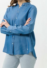 Jasnoniebieska koszula zapinana na guziki o miękkiej teksturze denimu, z długimi rękawami, zaokrąglonym końcem i kontrastującymi guzikami z przodu.