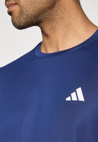 Tumši zila sportiska īsām piedurknēm krekls ar apaļu apkakli un teksturētu audumu. Kreisajā krūtīs ir balts trīsstūra logotips.