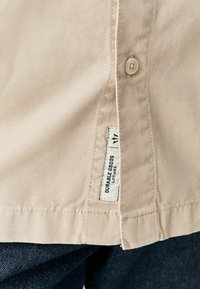Chemise en tissu beige avec un bouton, étiquette à l'ourlet portant "DURABLE GOODS" et "LOT 002," associée à un denim foncé. Texture lisse et coupe standard.