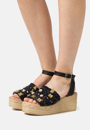 Sandales compensées noires en tissu tressé avec des accents dorés et de perles, dotées d'une sangle autour de la cheville et d'une semelle compensée enveloppée de jute.