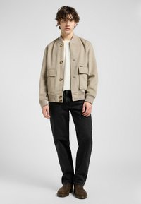 Beige ull bomberjakke med knappestolpe, to frontlommer, ribbede ermer, brukt over en hvit skjorte, kombinert med svarte jeans og brune støvler.