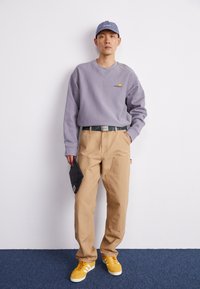 Sweatshirt gris à capuche avec logo, pantalon cargo beige, baskets jaunes et casquette bleue. Tient un objet matelassé gris foncé.