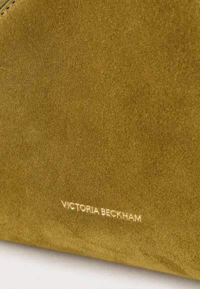 Κοντινό πλάνο υφάσματος από σουέτ ελιάς με κεντημένο το λογότυπο «Victoria Beckham» σε χρυσά γράμματα στο κάτω άκρο.