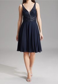 Laivaston mekko, jossa on syvä V-pääntie, koristeellinen vartalonosa, jossa on paljetteja, ja virtaava hame. Mallilla on päällään harmaat nauhaketjut.