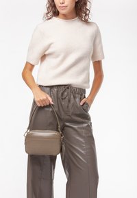 Piccola borsa rettangolare di colore marrone tenue, con una superficie tessuta e dettagli lisci, tenuta da una persona che indossa un maglione chiaro e pantaloni grigi.