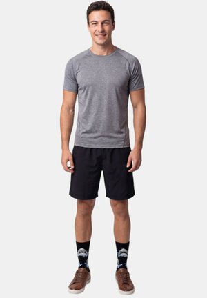 Hombre sonriente de pie con camiseta gris de manga corta, pantalones cortos negros, calcetines negros con diseño de tiburón y zapatos marrones sobre fondo blanco liso.