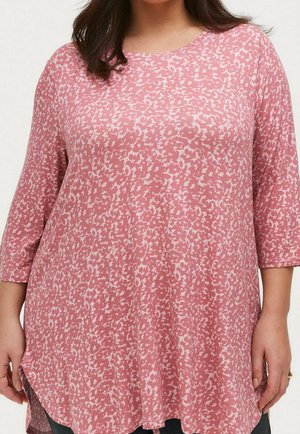 Woman wearing a loose, pink, floral-patterned tunic with three-quarter sleeves and rounded neckline, standing against a plain background.