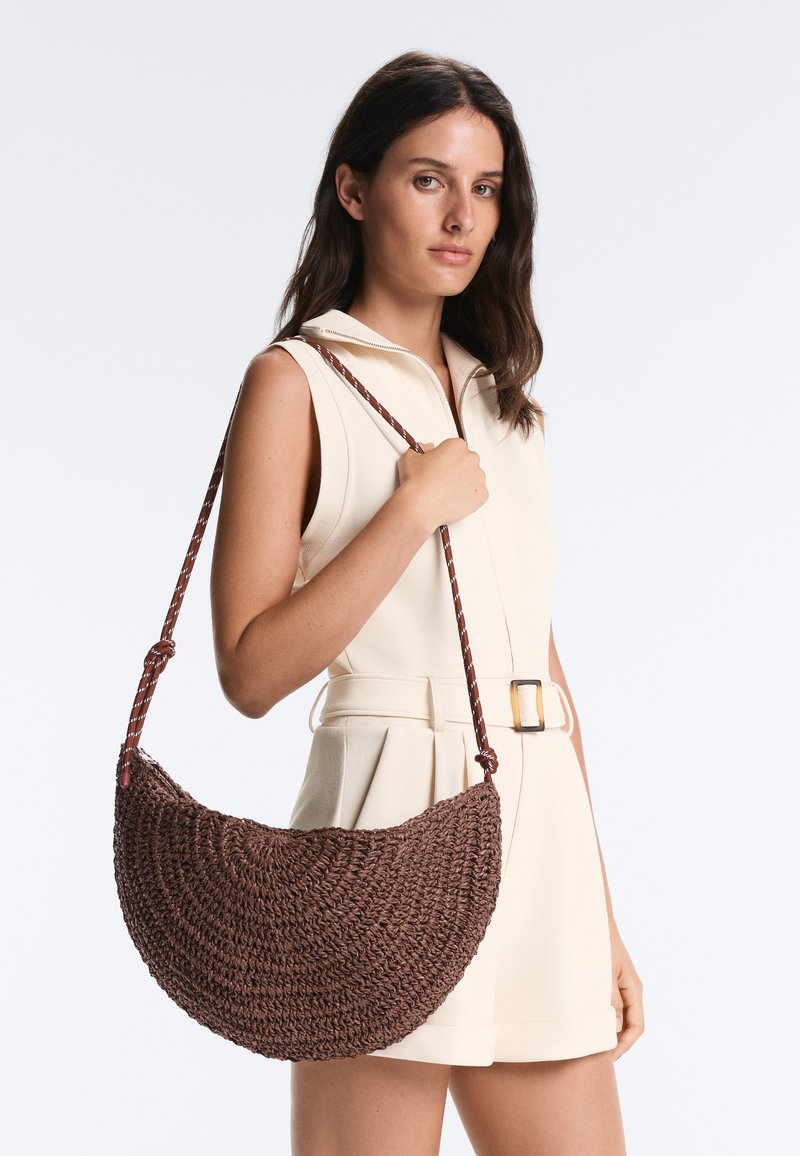 Femme en combinaison beige sans manches tenant un grand sac à bandoulière marron tissé avec une bandoulière tressée sur fond uni.